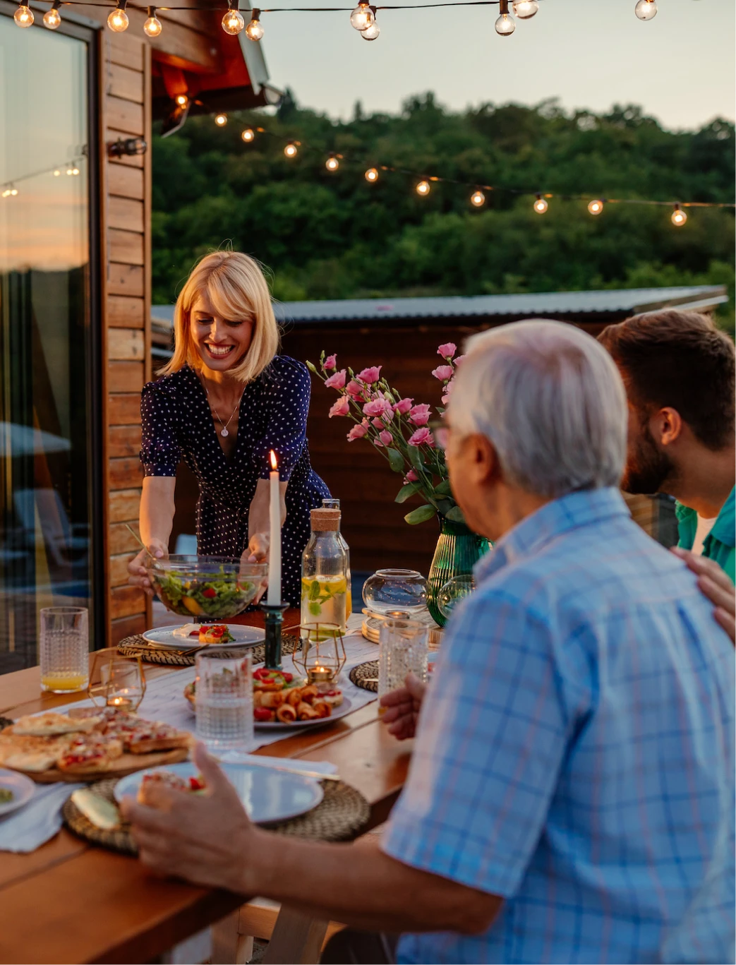 A group of people enjoying an outdoor meal at sunset. A woman is placing a salad bowl on the table, which is adorned with dishes, candles, and flowers. String lights add a warm ambiance to the cozy setting.