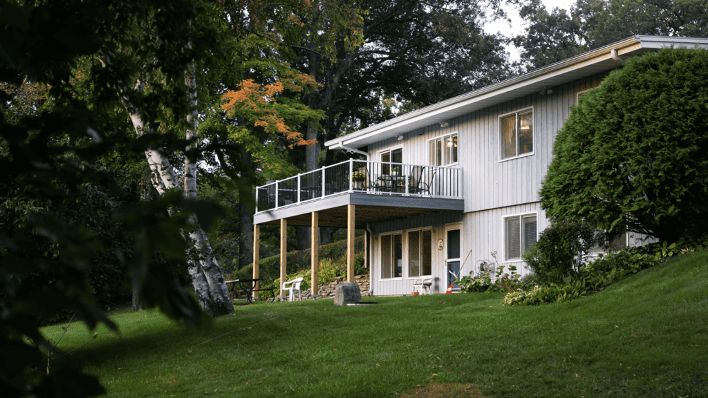 A two-story house with a raised deck and outdoor furniture, set on a grassy slope surrounded by lush trees. The upper structure is wooden with several windows, and the surrounding area includes a picnic table and a small patio.