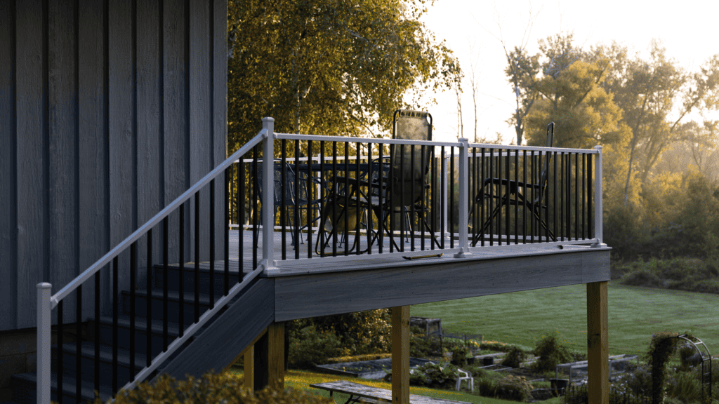 A deck with patio furniture and black railings is attached to a blue house, overlooking a lush green backyard with trees. The scene is lit by warm, early morning or late afternoon sunlight.