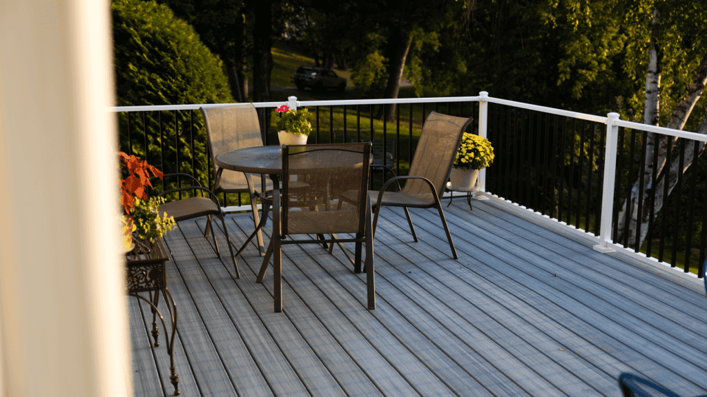 A sunlit patio features a round glass table with several mesh chairs. Flower pots with vibrant plants are positioned around the table on the wooden deck. A lush green lawn is visible in the background.