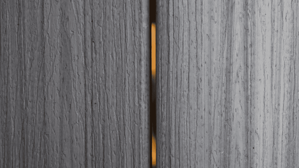 Close-up of two vertical wooden panels with a small gap between them. A sliver of warm light is visible through the gap, contrasting with the cool, textured gray surface of the wood.