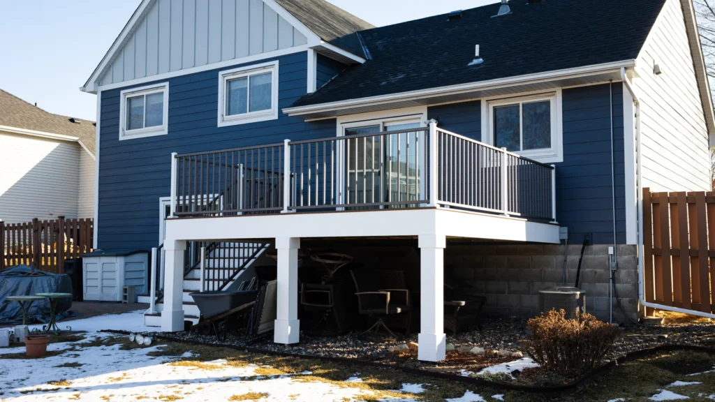 A blue two-story house with a raised porch featuring a metal railing. Beneath the porch are stacked chairs and other items. The yard has patches of snow and a small garden area. The fence and neighboring house are visible in the background.