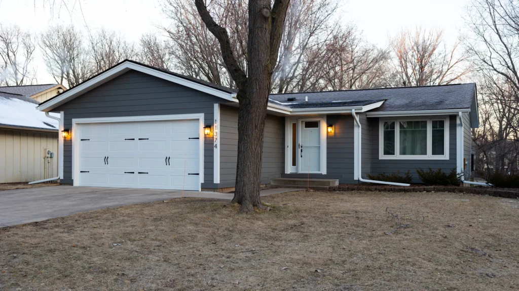 A single-story, gray house with a two-car garage and a tree in the front yard. The house has white trim and a concrete driveway. The grass in the yard is brown, and bare trees are visible in the background.