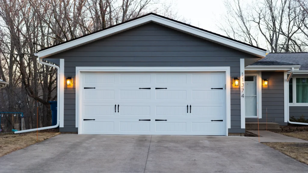 A gray house with a closed white garage door on a concrete driveway. The garage door has decorative black handles and lantern-style lights on either side. Leafless trees are visible in the background.