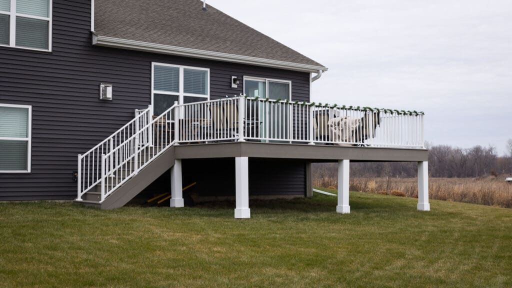A modern house with dark siding features a raised outdoor deck with a white railing. The grassy backyard extends to a distant, naturally vegetated area under an overcast sky. A set of stairs leads down from the deck.