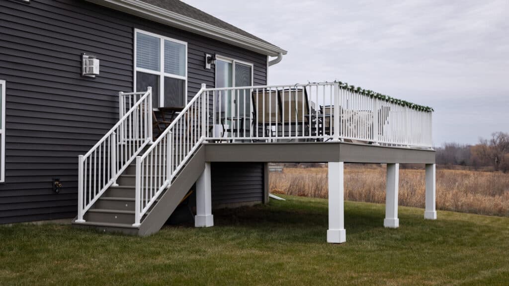 A house with a gray exterior features a raised deck with white railings. The deck has stairs leading down to a grassy yard. In the background, a field and cloudy sky are visible.