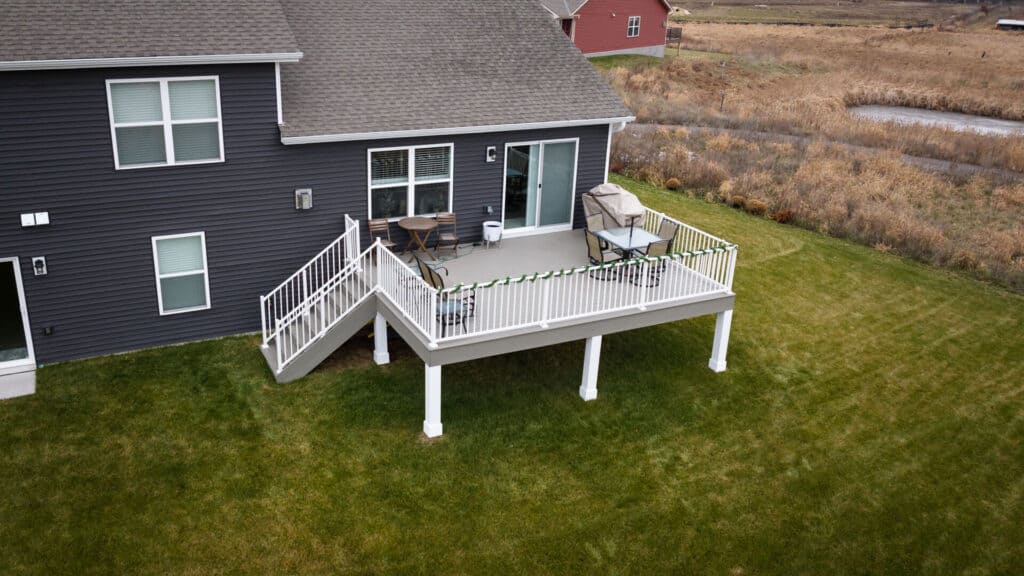 Aerial view of a house with a dark gray exterior and white-framed windows. The house features a white-railed, elevated deck with stairs leading down to a green lawn. The countryside landscape surrounds the property.