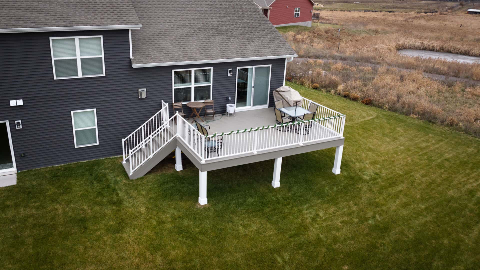 Aerial view of a house with a dark gray exterior and white-framed windows. The house features a white-railed, elevated deck with stairs leading down to a green lawn. The countryside landscape surrounds the property.