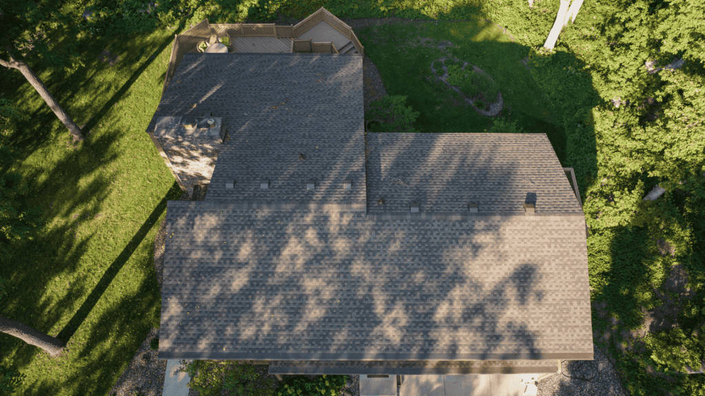 Aerial view of a house with a brown shingle roof surrounded by green trees. Shadows from the trees are cast on the roof and the surrounding grassy area. The house has a few architectural elements and a driveway visible at the bottom.
