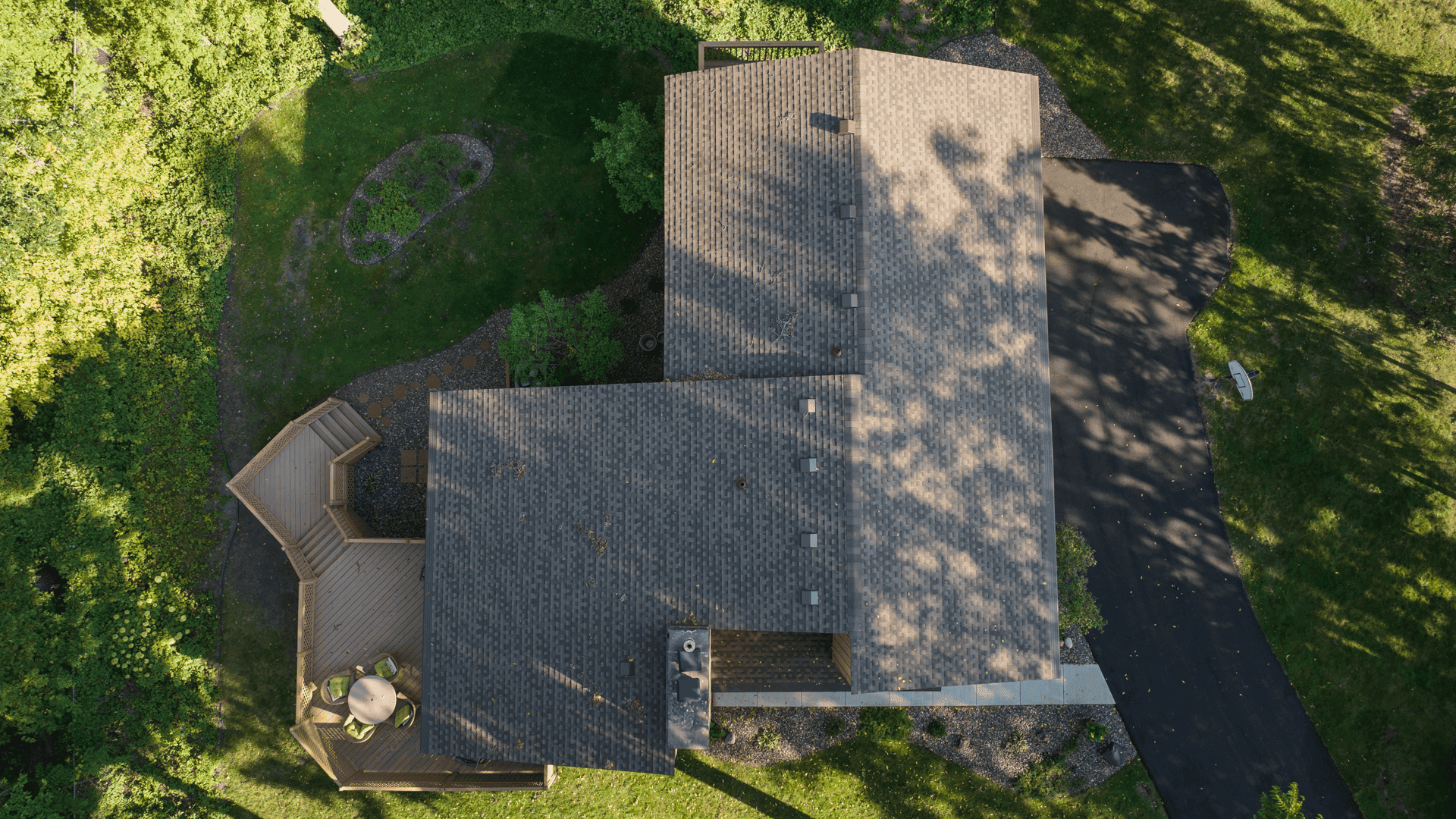Aerial view of a house with a dark gray roof, surrounded by lush green trees. The property includes a wooden deck with outdoor furniture and a black driveway. Shadows from the trees create patterns on the roof and lawn.