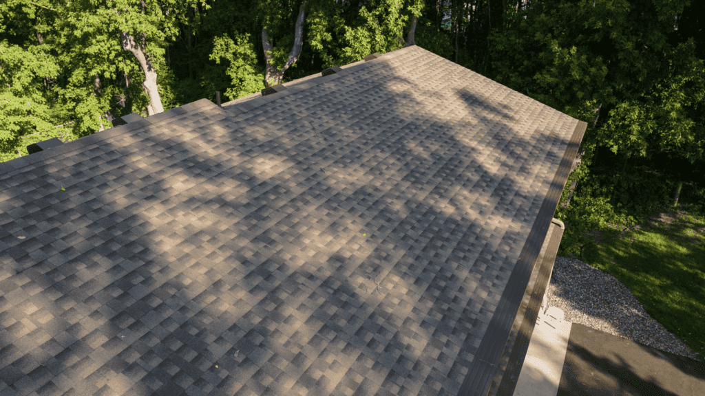 Aerial view of a house roof covered with brown asphalt shingles. The roof is surrounded by dense green trees, casting shadows on its surface. The shingles form a grid-like pattern of various shades of brown.