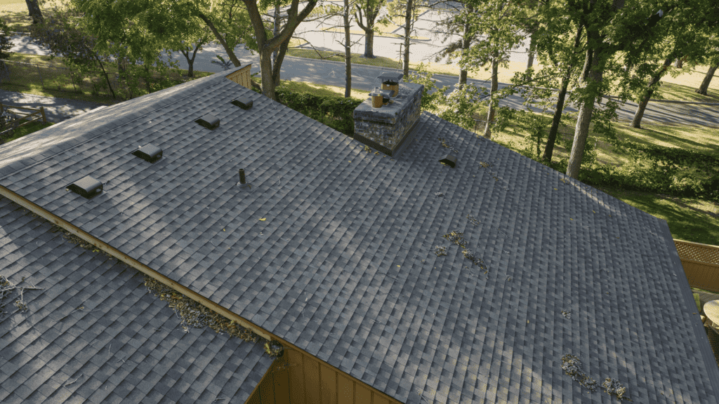 A rooftop view of a house surrounded by trees. The roof is covered with gray shingles and has a brick chimney. Some fallen leaves and twigs are scattered across the shingles. The background shows a road lined with trees.