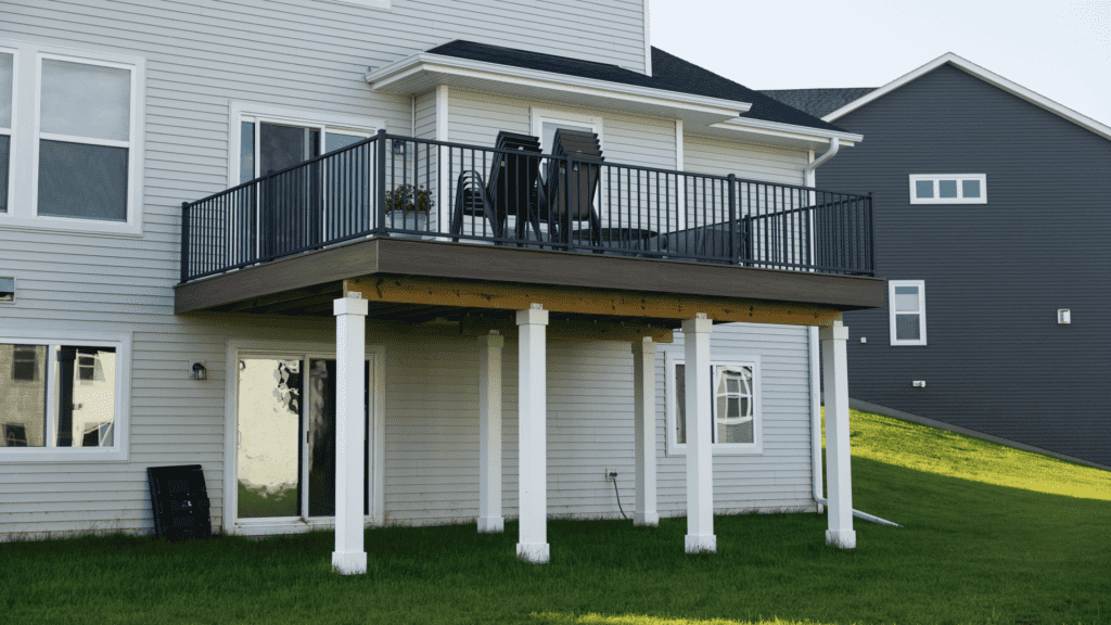 A modern two-story house with light grey siding features a covered patio supported by white pillars. The patio has a black metal railing and is furnished with stacked chairs. The house sits on a well-manicured green lawn.