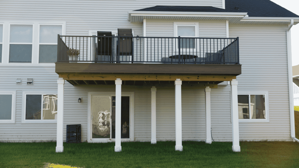 A two-story house with a wooden deck supported by white posts. The deck has a black metal railing and outdoor furniture, including a table with chairs. Below the deck is a glass door and windows, opening to a grassy yard.