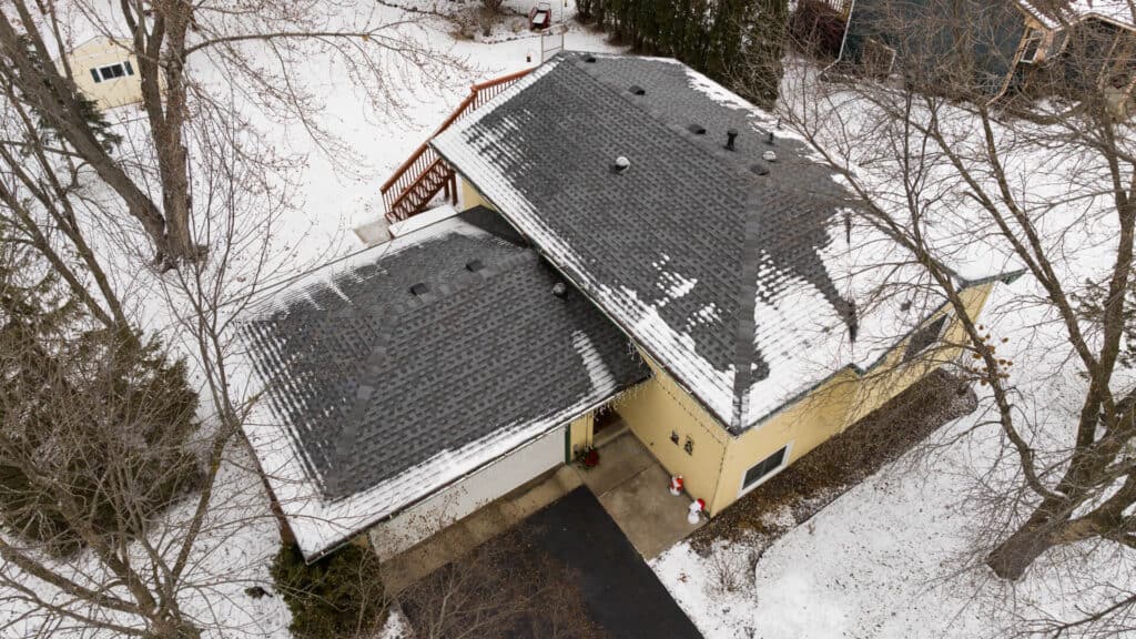 Aerial view of a house with a partially snow-covered shingle roof. Snow patches are visible around the area, and bare trees surround the house. A driveway leads to the garage, and the ground is lightly dusted with snow.