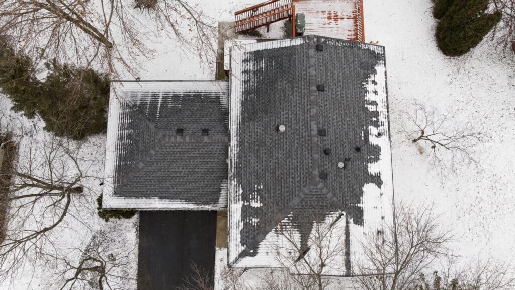 Aerial view of a snow-covered house with a dark roof, surrounded by leafless trees. The driveway is partially visible, leading to the road. A red wooden deck is attached to the back of the house, creating a contrast with the white snow.
