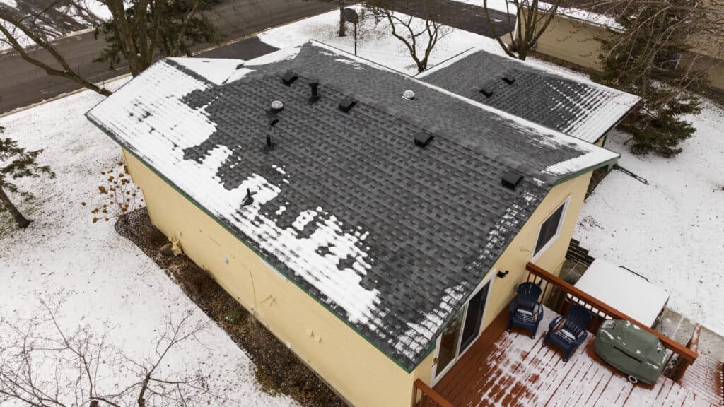 Aerial view of a yellow house with a snow-covered, dark shingled roof. There's a wooden deck with chairs and a covered grill. The ground and surrounding trees are covered in light snow, indicating a winter setting.