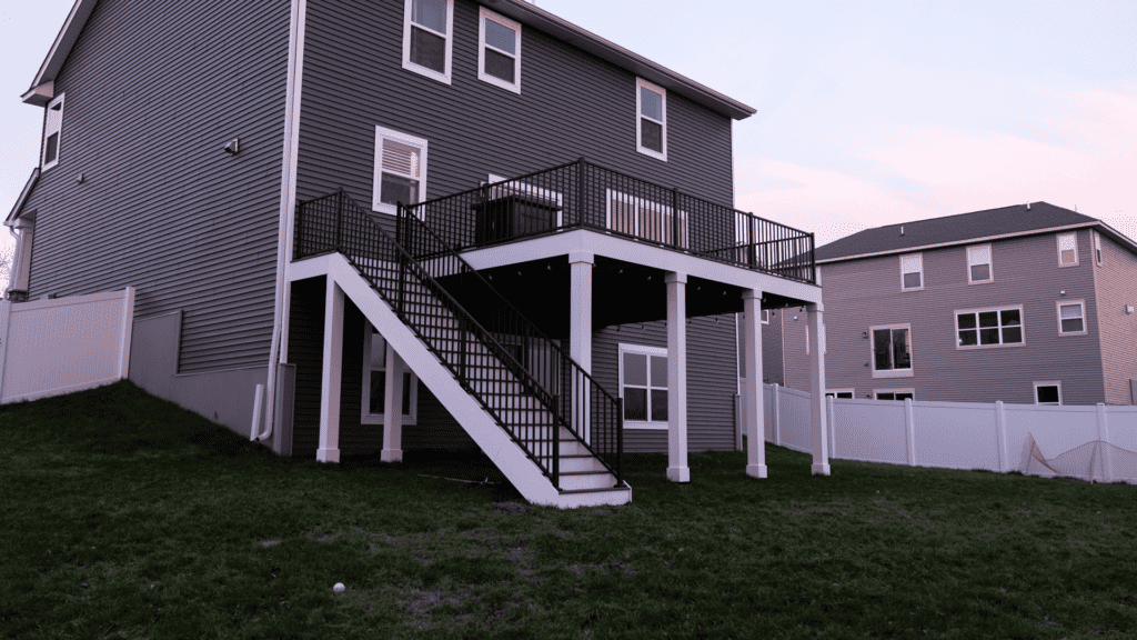 A two-story house with blue-gray siding and a white foundation features a wooden deck with black railings and stairs leading down to a grassy backyard. A white fence encloses the yard, with another similar house visible in the background.