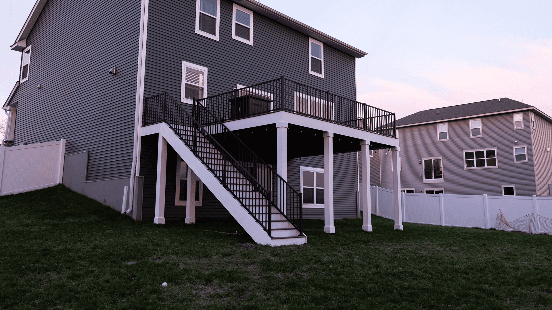 A two-story house with blue-gray siding and a white foundation features a wooden deck with black railings and stairs leading down to a grassy backyard. A white fence encloses the yard, with another similar house visible in the background.