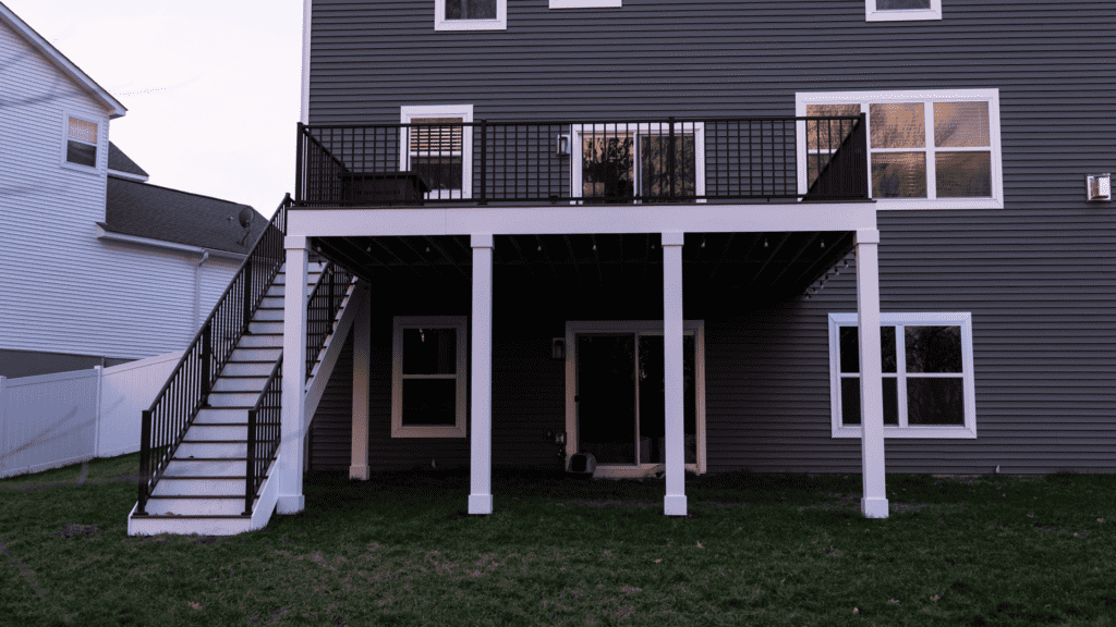 A two-story house with a gray exterior, featuring a raised wooden deck on the second floor. The deck has a black railing and a staircase leading to the grassy backyard below. Windows adorn both the upper and lower floors.