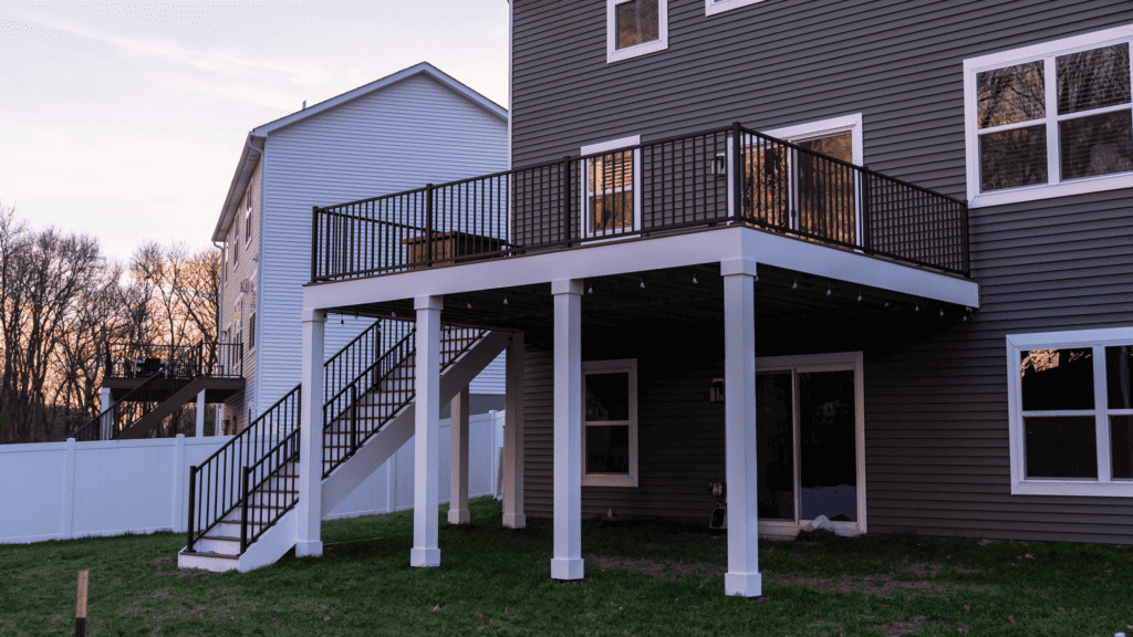 A two-story house with gray siding features an elevated wooden deck with black railings and stairs leading down to a grassy backyard. The white fence borders the property, and another house is visible nearby.