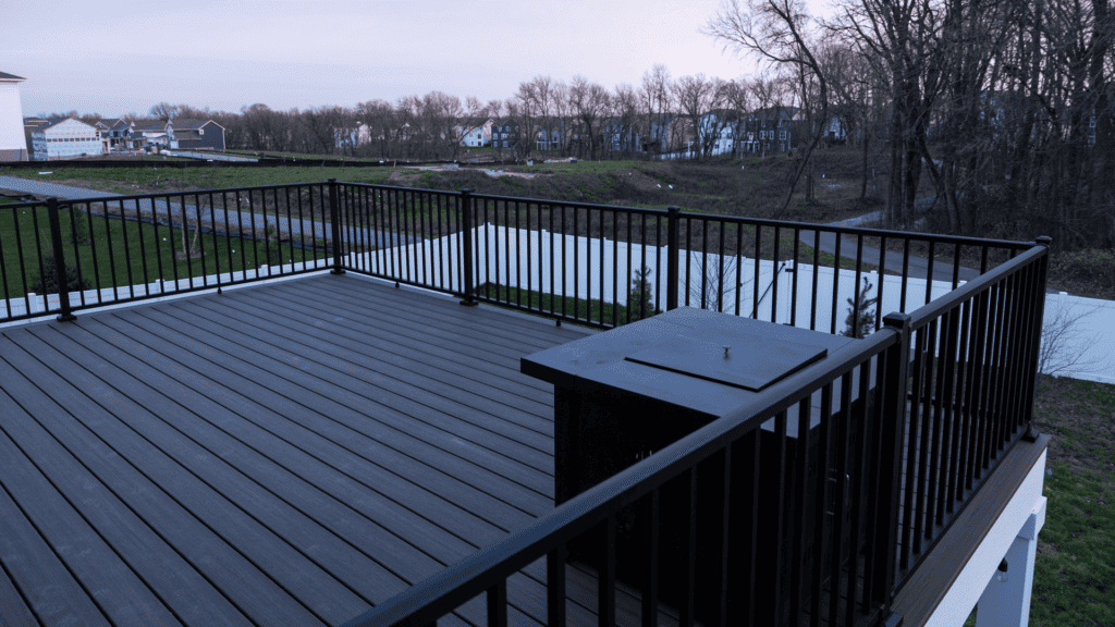 A spacious rooftop deck with dark railings overlooks a suburban neighborhood at dusk. Trees and houses line the background, and a small grill is placed in one corner of the deck. The atmosphere is peaceful and serene.