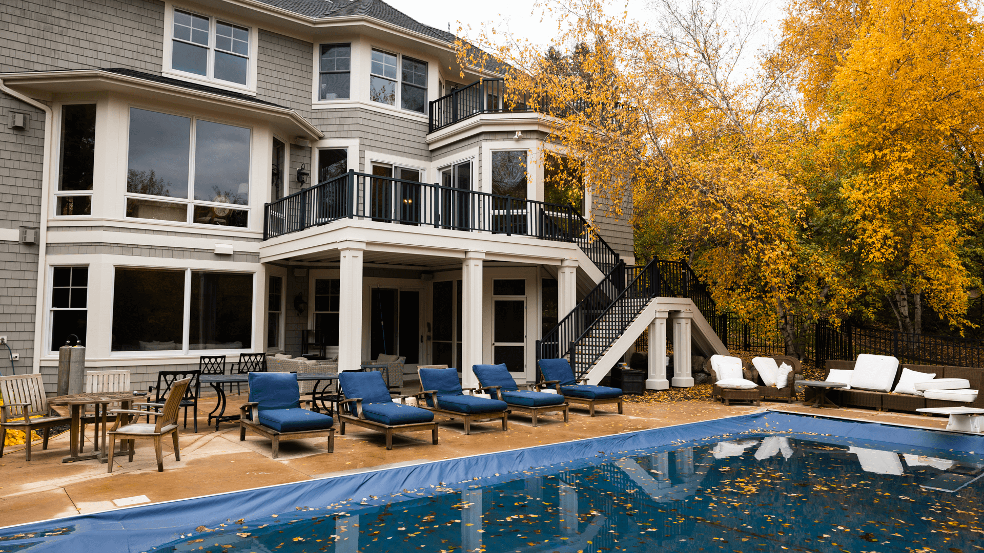 A large house with gray siding and multiple windows is surrounded by autumn trees. In the backyard, there's an outdoor pool with a cover, and several blue lounge chairs and tables on the patio.