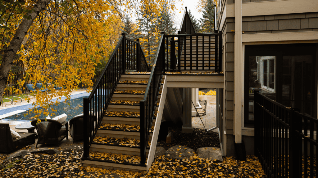 Outdoor scene with a staircase covered in fallen yellow leaves next to a small patio with chairs by a pool. Trees with autumn foliage surround the area, adding a warm, seasonal feel to the setting.