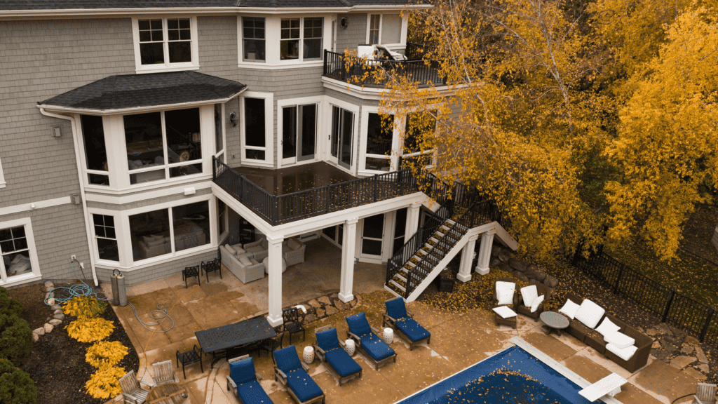 Aerial view of a large gray house with bay windows and multiple balconies. The backyard features a swimming pool surrounded by lounge chairs and a patio area. Trees with autumn foliage frame the scene, adding vibrant yellow-orange accents.