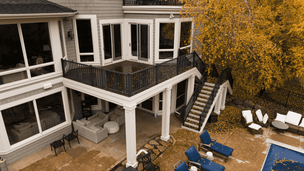 Aerial view of a two-story house with large windows, a patio, and a balcony. There's a pool surrounded by blue lounge chairs in the backyard. Autumn leaves cover the ground and nearby trees, adding a yellow hue to the scene.
