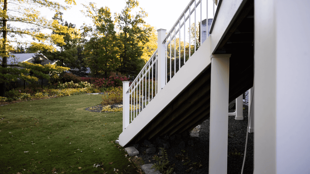 A white staircase with railings leads from a wooden deck to a grassy yard. The yard is lined with trees and shrubs, and the scene is warmly lit by the sun.
