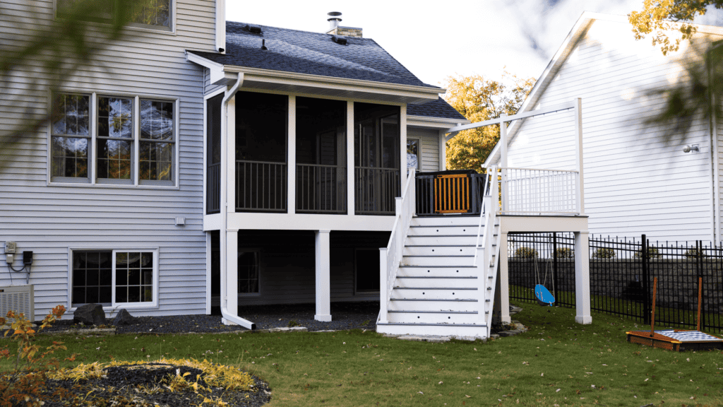 A two-story house with gray siding features a screened porch on the second level, accessible by a white staircase. A small fenced yard has green grass, trees, and a game set on the ground. Nearby, a blue swing hangs from a tree.