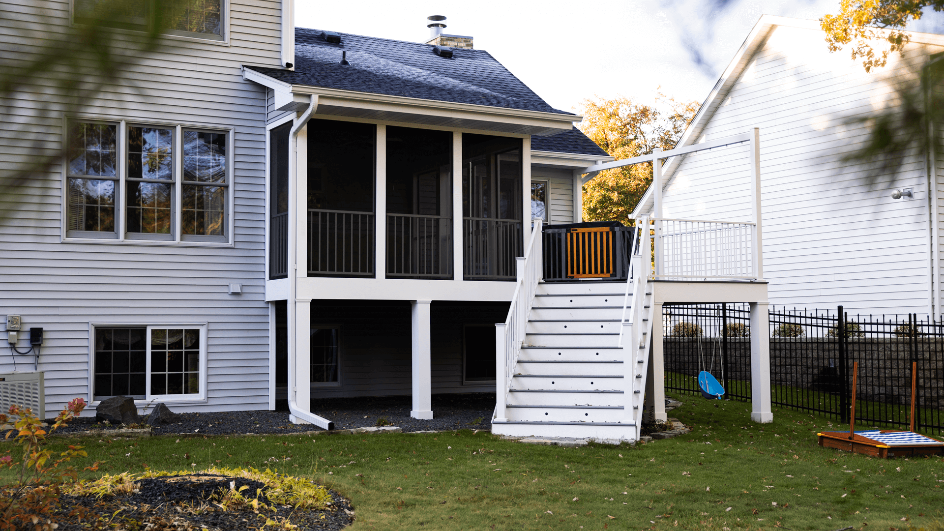 A two-story house with gray siding features a screened porch on the second level, accessible by a white staircase. A small fenced yard has green grass, trees, and a game set on the ground. Nearby, a blue swing hangs from a tree.