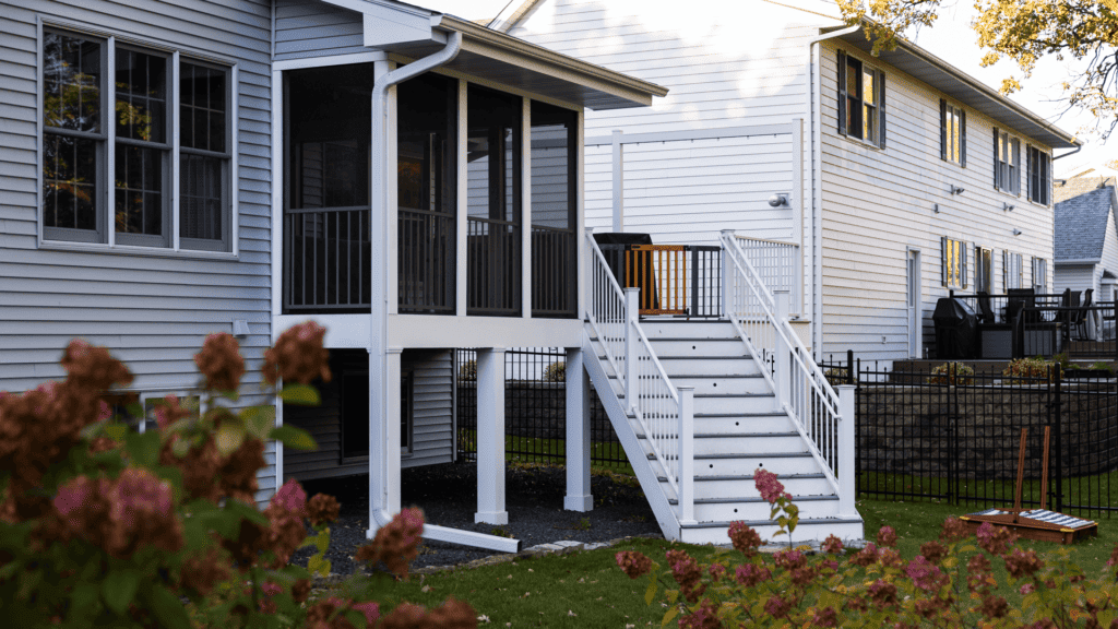 A suburban house features a screened-in porch elevated on white pillars, accessible by a staircase with black railings. The yard includes a green lawn and flowering plants, with a neighboring house and wooden fence in the background.