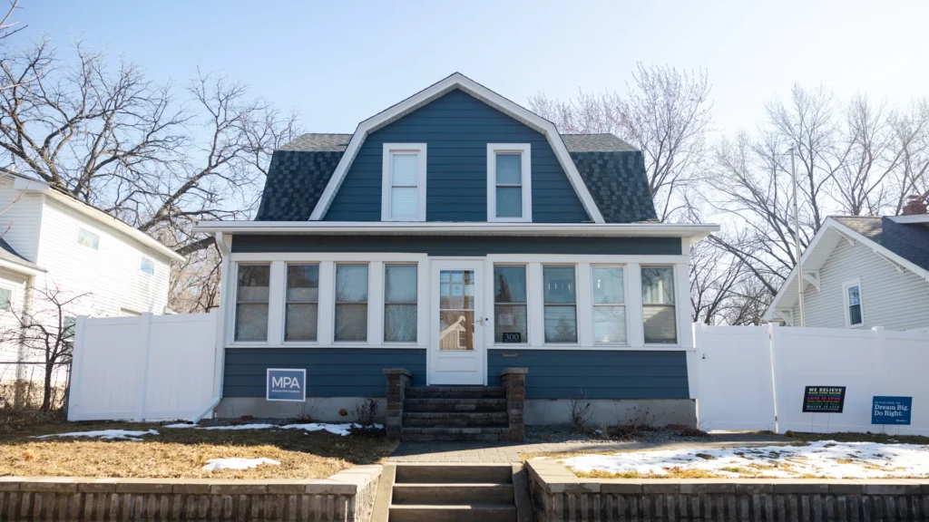 A two-story blue house with a steep roof is surrounded by a white fence. The front yard has a small amount of snow and some campaign signs. Leafless trees are visible in the background under a clear sky.