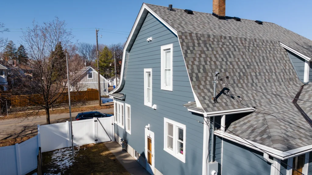 A two-story blue house with white trim features a steep grey shingled roof and a visible chimney. The surrounding yard has partial grass and bare trees, with a white fence and a street in the background under a clear blue sky.