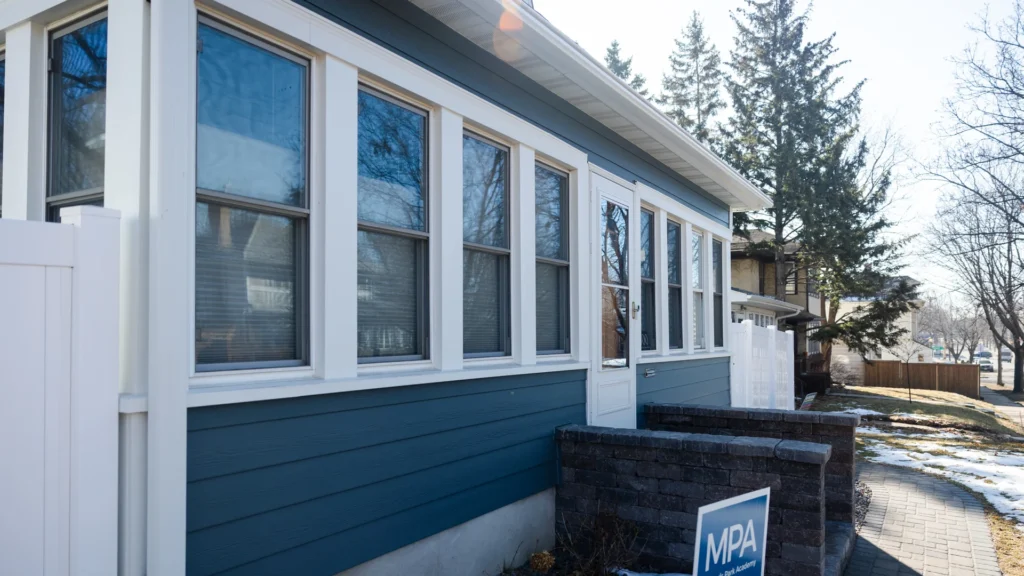 A blue house with large windows and a white door is shown from the side. The front features a small brick wall and a "For Sale" sign. Trees and a driveway are visible in the background on a sunny day.
