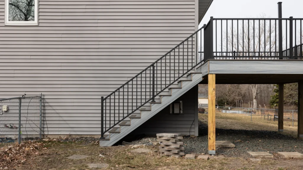 A gray, two-story house features a second-floor deck with black metal railings. Stairs lead down to a grassy yard with a stack of concrete blocks nearby. The surrounding area is fenced, and bare trees are visible in the background.