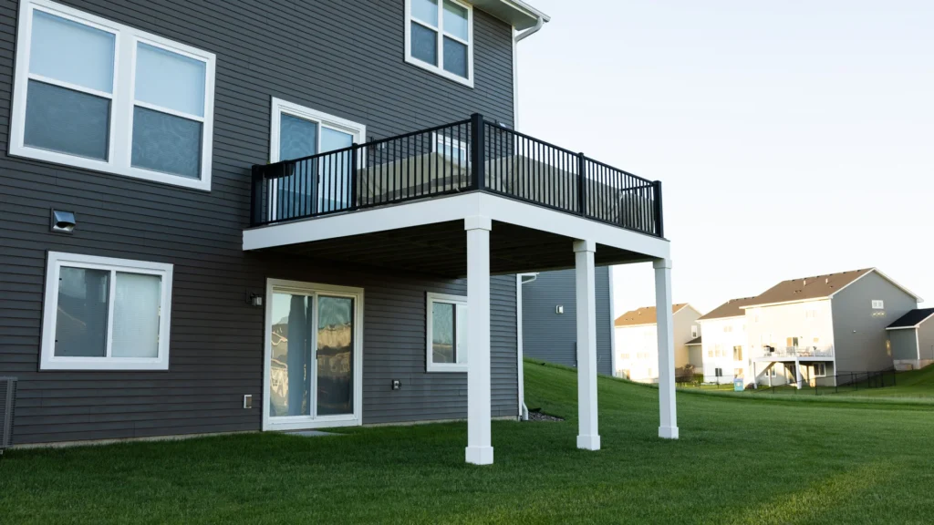 A two-story house with gray siding features a raised deck with black railings supported by white columns. A grassy lawn surrounds the house, and neighboring houses are visible in the background under a clear sky.
