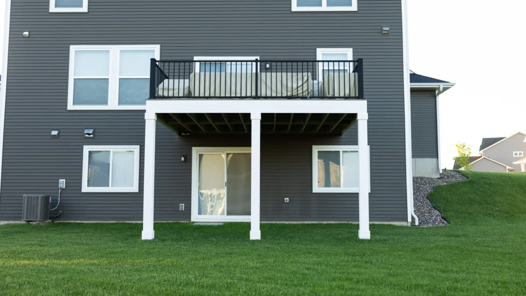 A two-story building with gray siding, featuring a small patio deck with white columns. The patio has a metal railing and outdoor seating. Below, there’s a sliding glass door leading to the backyard with well-maintained green grass.