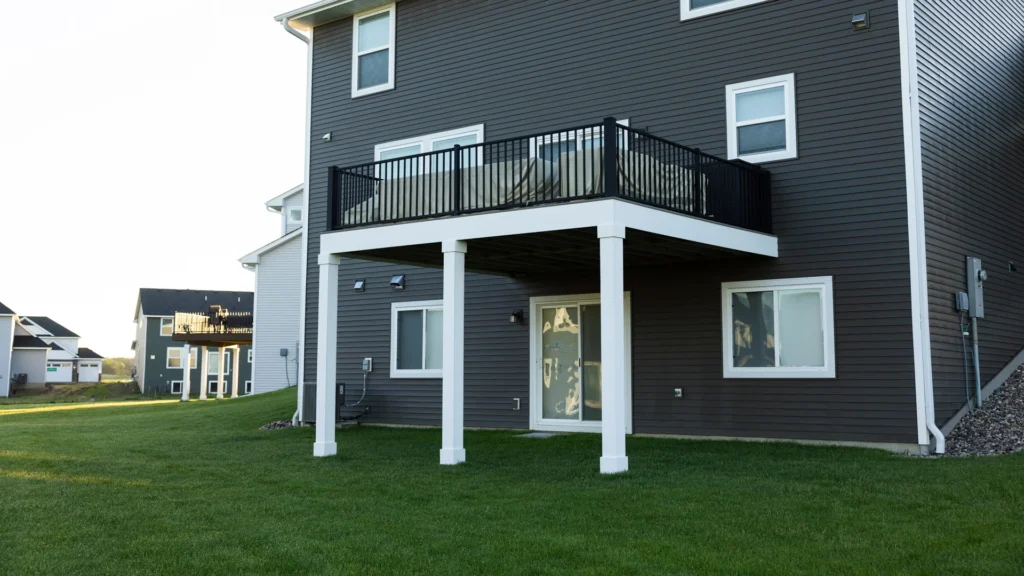 A modern gray two-story house with a raised balcony supported by white pillars. The balcony has a black railing, and there is a glass sliding door on the ground floor. The house is surrounded by a well-maintained grassy lawn.