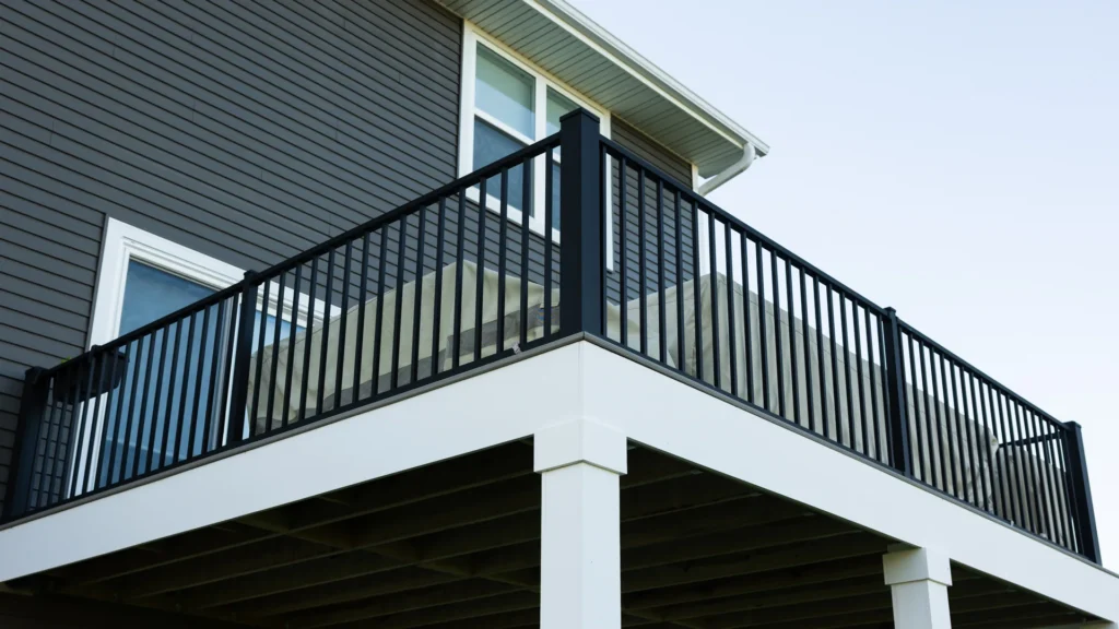 A modern two-story house with a gray exterior features a raised deck with black metal railings. The deck is supported by white posts and is adjacent to a window on the upper floor. The clear sky is visible in the background.