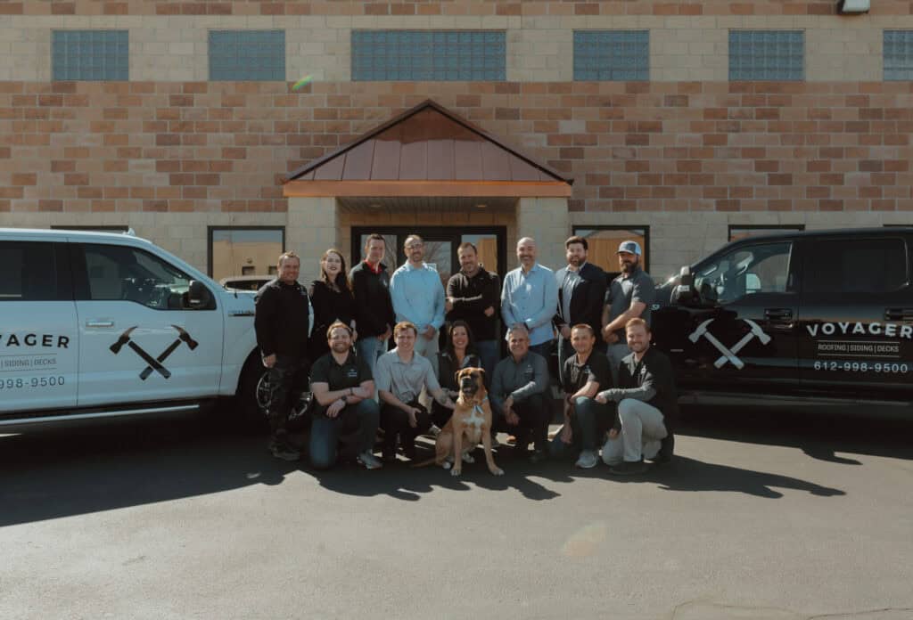 A group of people are posing for a photo in front of a brick building. Two black vans with "Voyager" written on them flank the group. A large dog sits at the front center, and the sky is clear and sunny.