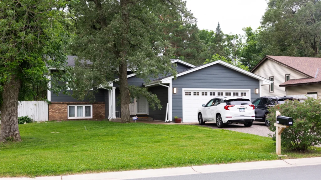 A suburban house with dark blue siding and a white garage door. A white SUV is parked in the driveway, and a lawn with trees surrounds the home. A mailbox is near the curb, and neighboring houses are visible.
