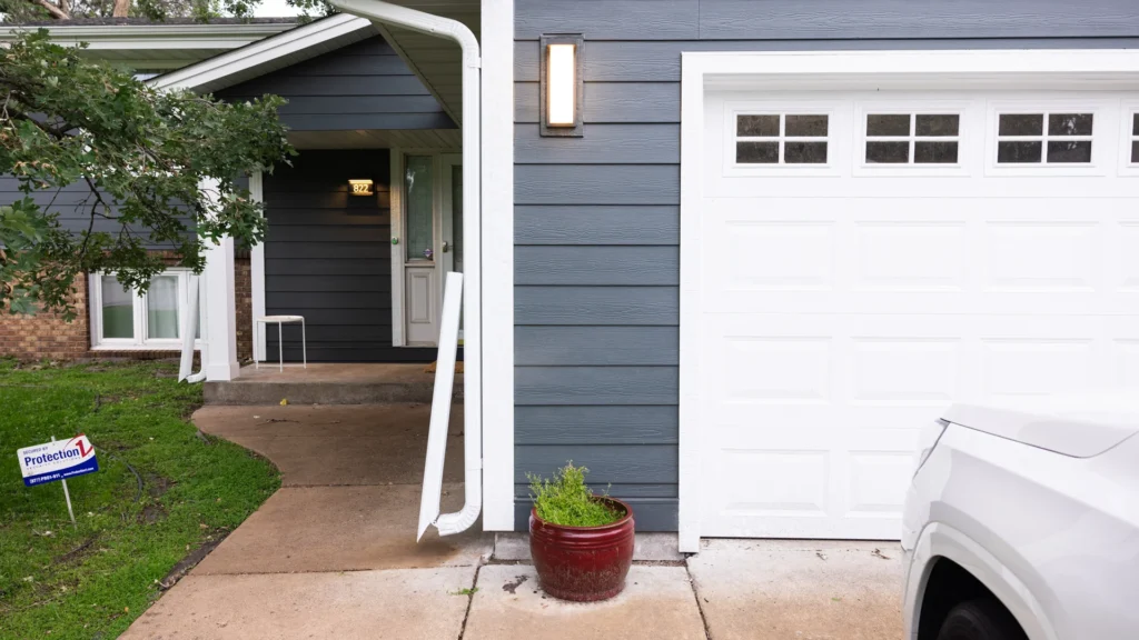 A suburban house with gray siding, a white garage door, and a small porch visible. There is a light above the porch and a potted plant near the garage. A sign with "Protection" is seen on the lawn alongside a portion of a white vehicle.