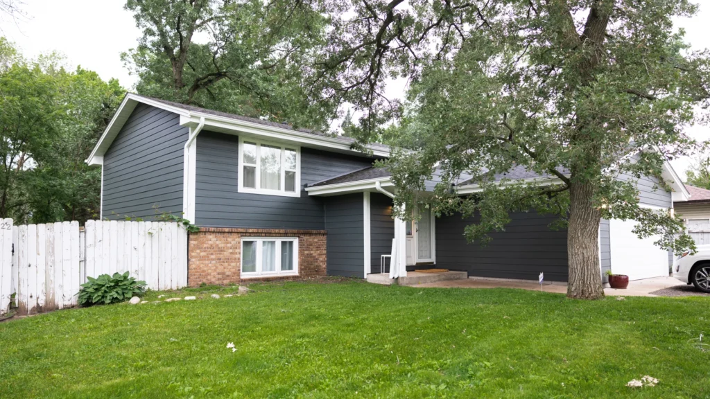 A two-story house with dark gray siding and a brick lower section, surrounded by tall trees and green lawn. A white picket fence is on the left, and a driveway with a car is visible on the right. A small porch is at the front entrance.