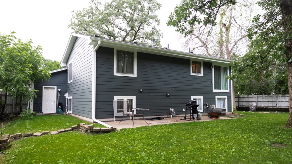 A two-story gray house with white trim, featuring a patio in the backyard. The patio has a table, chairs, and a grill. Green grass and trees surround the area, with a partial view of a white fence in the background.