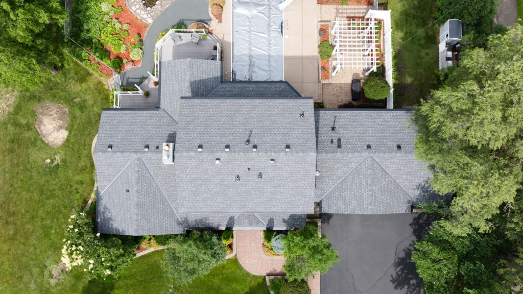 Aerial view of a large house with a gray roof, surrounded by landscaped gardens and a paved driveway. A patio and a white pergola are visible in the yard, along with green trees and shrubs around the property.