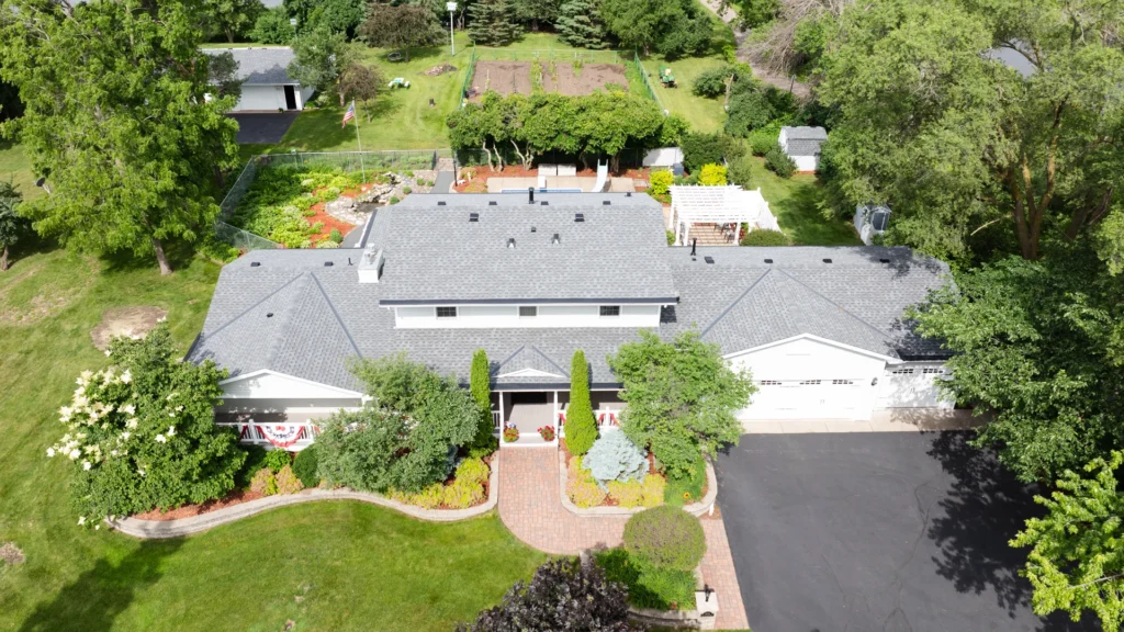 Aerial view of a large, gray-roofed house surrounded by lush greenery and gardens. A driveway leads to a multi-car garage, and there is a brick path lined with vibrant landscaping, including trees and shrubs. A fenced garden area is visible in the background.