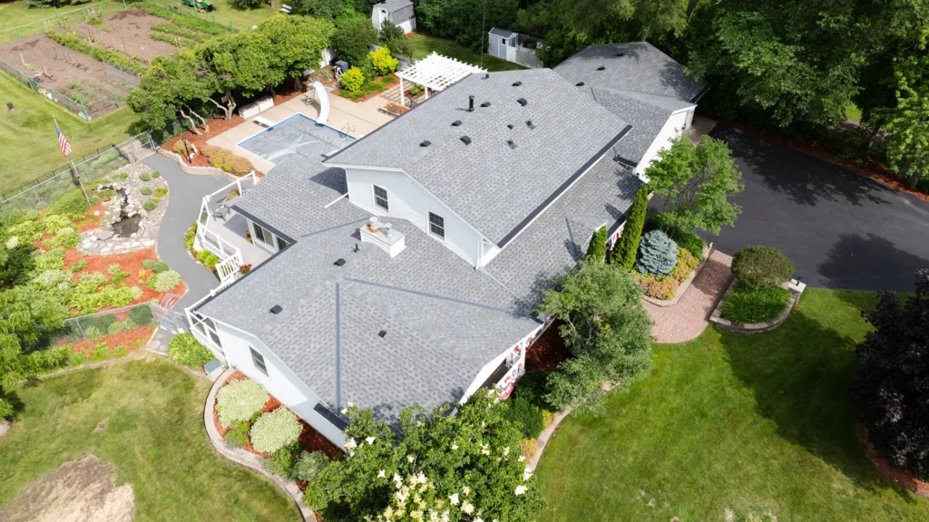 Aerial view of a large grey-roofed house with a landscaped garden. The property has surrounding greenery, a driveway, and a paved patio area. A flagpole and vegetable garden are visible on the left.